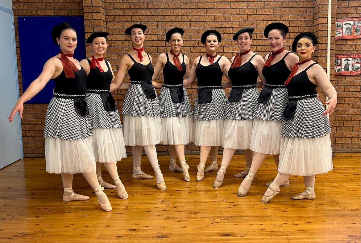 A group of young women dressed in ballet performance outfits - they're wearing berets, red neckerchiefs, black singlets, flowing skirts and pointe shoes.